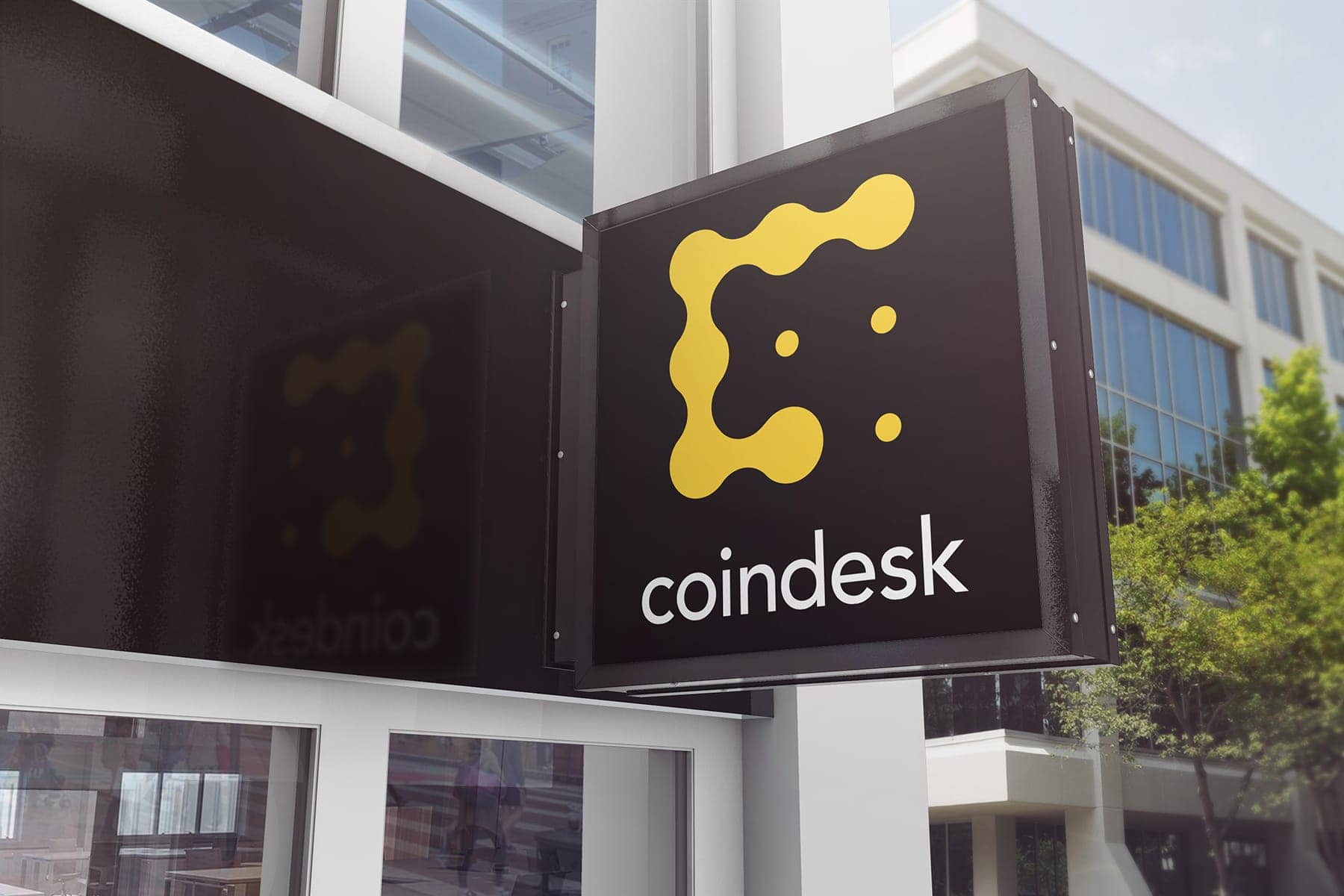 A projecting building sign in near-black carrying the yellow CoinDesk mark above the lowercase wordmark, mounted on a glass and white stone facade.